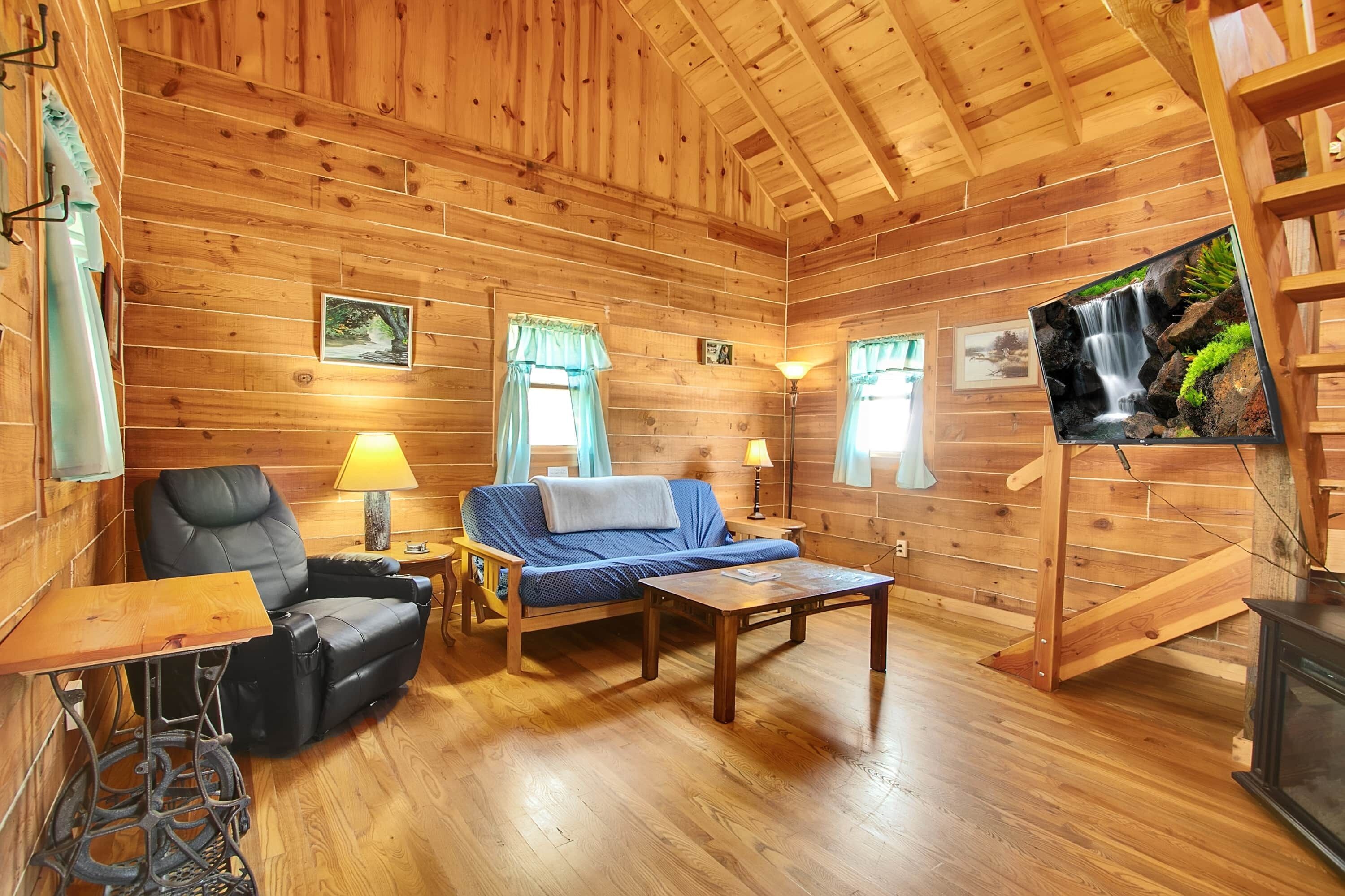 Ein Holzblockhaus im Inneren, mit einer Couch, einem Sessel, einem Tisch und einem an der Wand befestigten Fernseher.