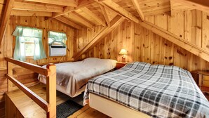 Une cabane en bois avec une chambre à coucher située en mezzanine, une fenêtre avec des rideaux et une table de chevet avec une lampe.