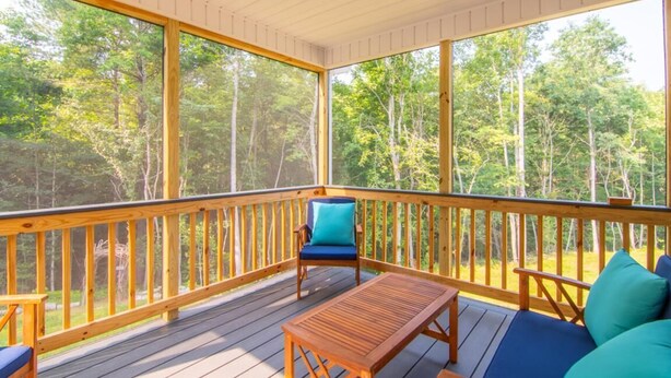 A covered outdoor deck with wooden railings, a blue cushioned chair, a wooden table, and a sofa with teal cushions. - Bluebird Cabin | WiFi & River Access (Elkin)