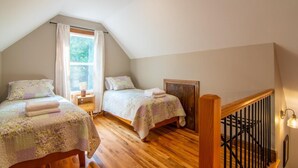 A bedroom with two beds, wooden flooring, a window with curtains, a nightstand, and a staircase with a railing. - Bluebird Cabin | WiFi & River Access (Elkin)