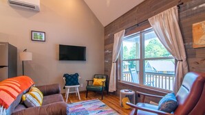 A cozy living room with a brown sofa, a colorful rug, and a wooden deck visible through the window. - Bluebird Cabin | WiFi & River Access (Elkin)