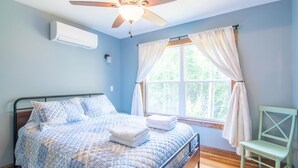 Un dormitorio con cama, ventilador de techo, aire acondicionado, ventana con cortinas y una silla.