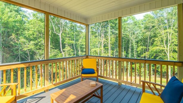 Una terraza cubierta al aire libre con barandas de madera, una silla amarilla con cojín azul y una mesa de madera.