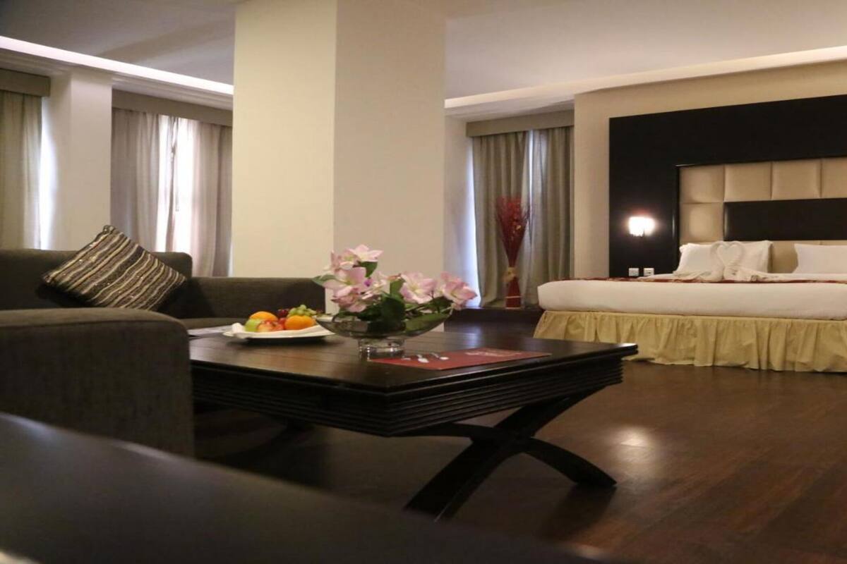 A hotel room with a bed, a sofa, a coffee table with fruit and flowers, and a nightstand with a lamp.