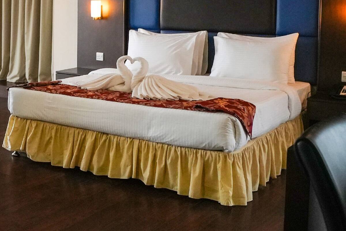 A hotel room with a large bed, a headboard with a blue panel, a wall-mounted light, and a curtain on the window.