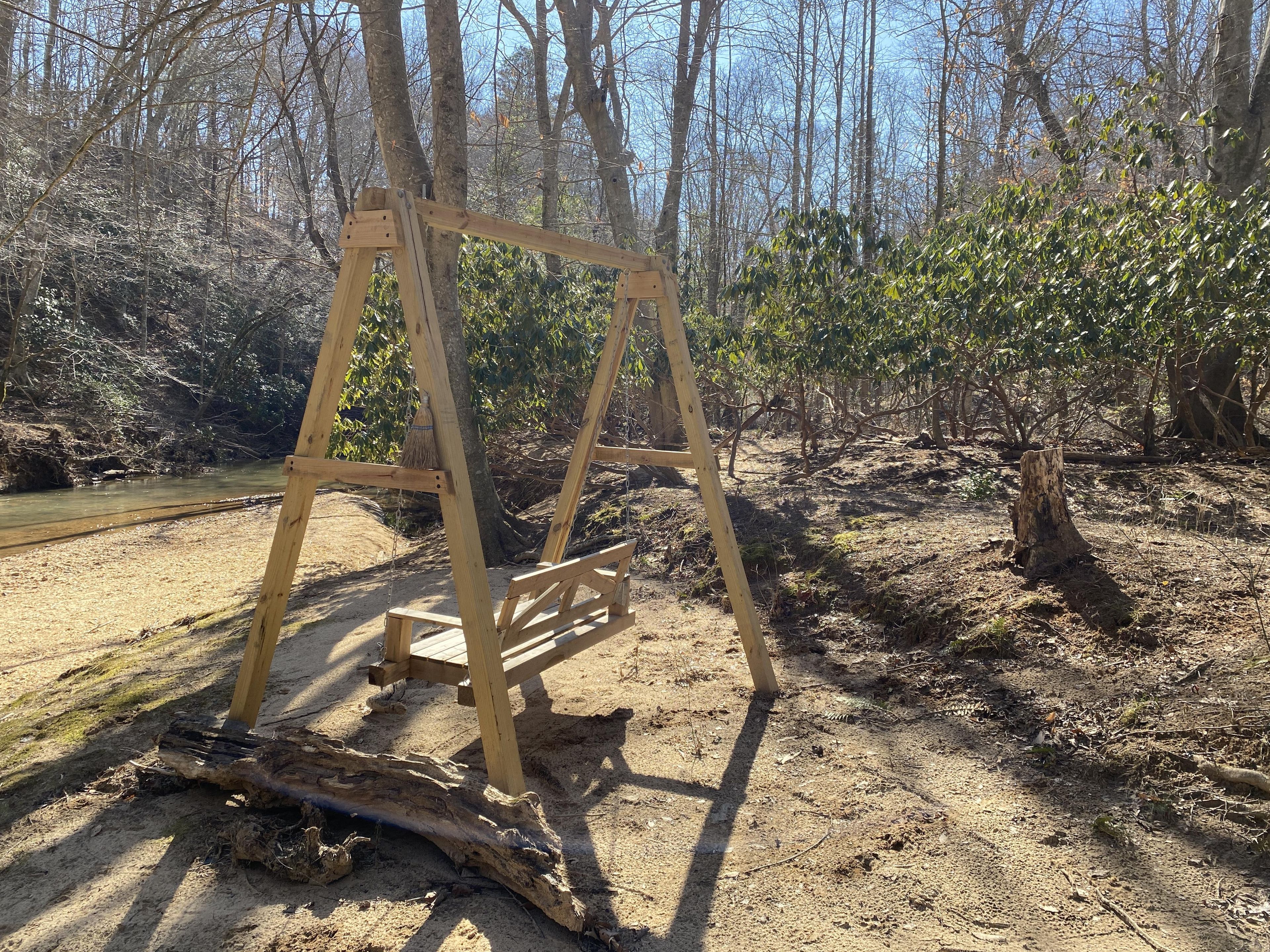 A wooden swing set in a forested area with a dirt path and a small stream.