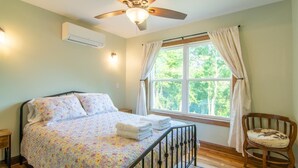 A bedroom with a bed, a ceiling fan, an air conditioning unit, a window with curtains, and a chair. - Monarch Cabin | WiFi & River Access (Elkin)