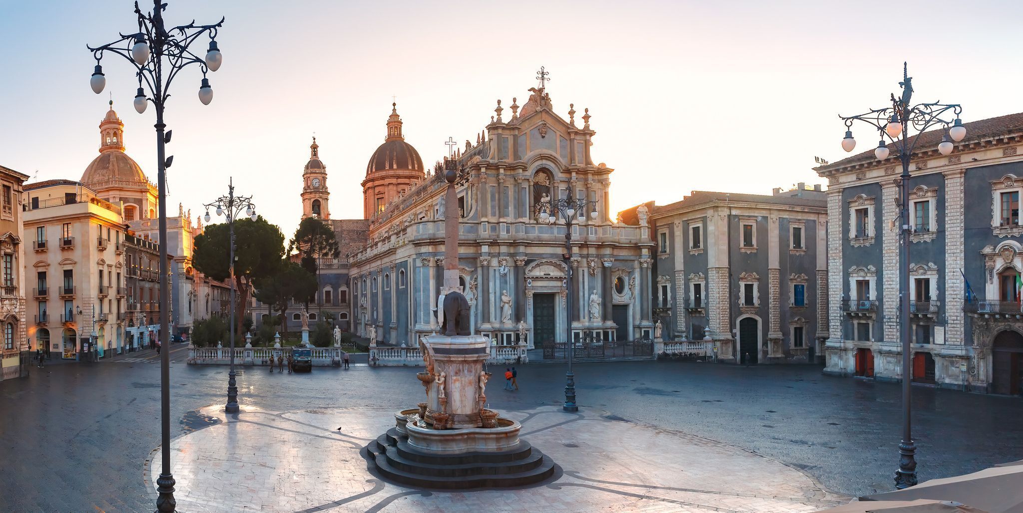 A historic plaza with ornate buildings, a central fountain, and a statue.