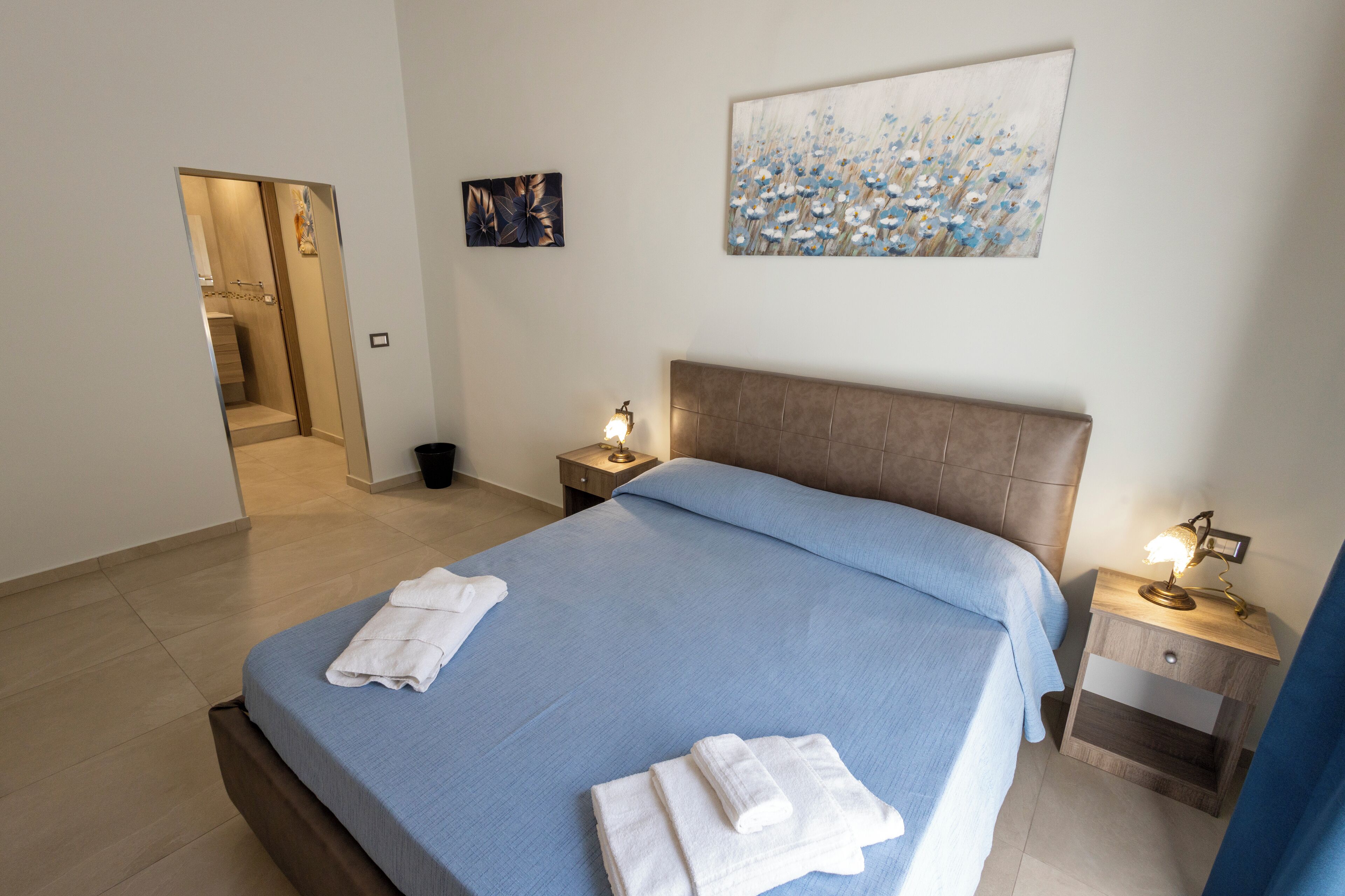 A bedroom with a bed, two bedside tables, a nightstand, a wall-mounted painting, and a door leading to a bathroom.