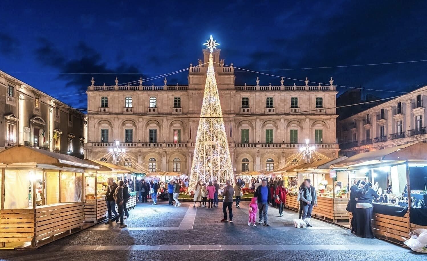 Un grand sapin de Noël illuminé se dresse devant un bâtiment historique doté de nombreux balcons, et le sol est mouillé, ce qui laisse supposer qu’il a plu récemment.