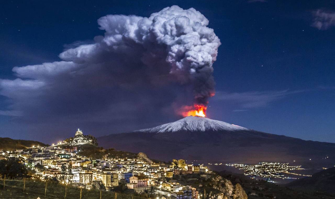 A volcano with a large plume of smoke and lava at the summit, a town with illuminated buildings in the foreground, and a mountain range in the background.