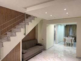 Un interno moderno con una scala, un divano, un'area pranzo con tavolo e sedie, e una cucina visibile attraverso una porta aperta.