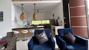 Ein modernes Wohnzimmer mit zwei blauen Sesseln, einer Holzbodenverkleidung, einer Küchenzeile mit Bar und einer großen Schiebetür.