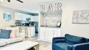 Ein modernes Wohnzimmer mit einer Couch, einem Couchtisch und einem blauen Sessel. Im Hintergrund ist eine Küche mit weißen Schränken und einer Ziegelrückwand zu sehen.