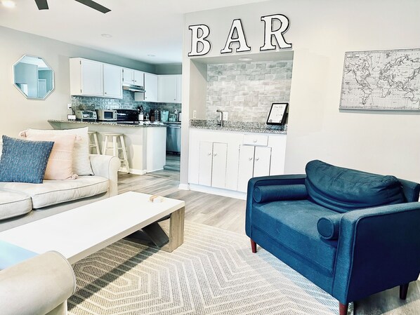 Una sala moderna con un sofá, una mesa de centro y un sillón azul. Se ve una cocina con armarios blancos y un revoque de ladrillo al fondo.