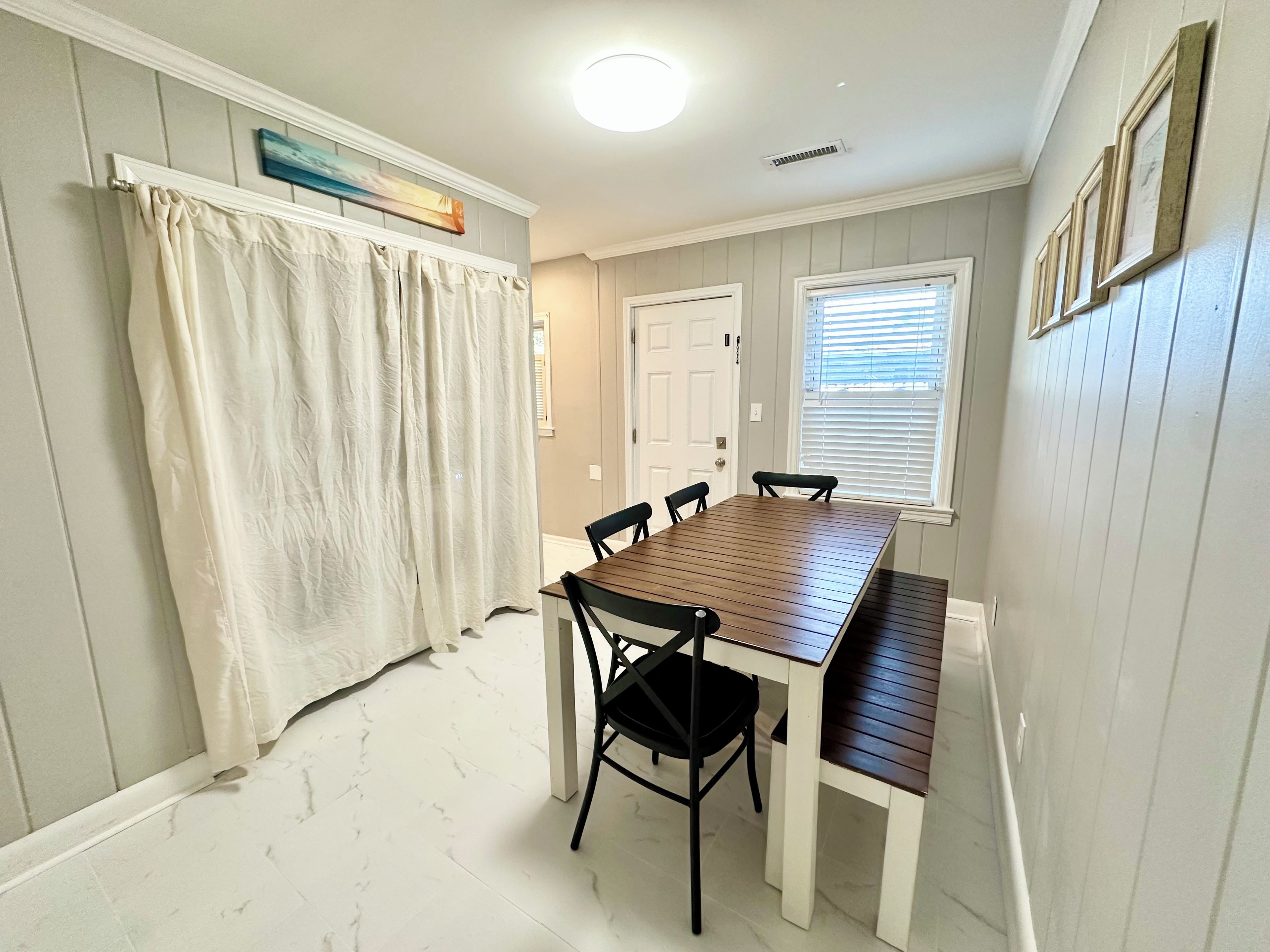 Un coin repas avec une table et des chaises en bois, un rideau blanc, une porte et une fenêtre avec des stores.