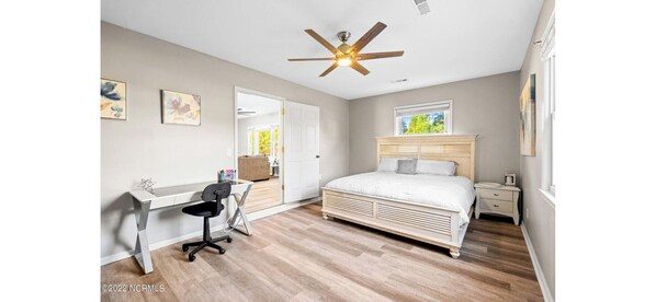 A bedroom with a bed, desk, chair, ceiling fan, and artwork on the walls.