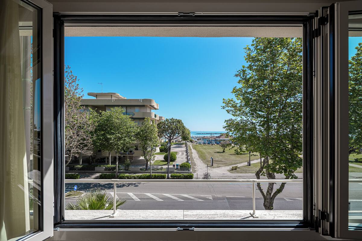 Vista desde una ventana con vista a una calle, un edificio moderno, árboles y cielo despejado.