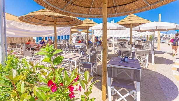 Un espace extérieur aménagé avec des tables et des chaises, protégé par des parasols en chaume, et entouré de tentes blanches et de chaises de plage.