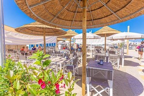 Un espace extérieur aménagé avec des tables et des chaises, protégé par des parasols en chaume, et entouré de tentes blanches et de chaises de plage.