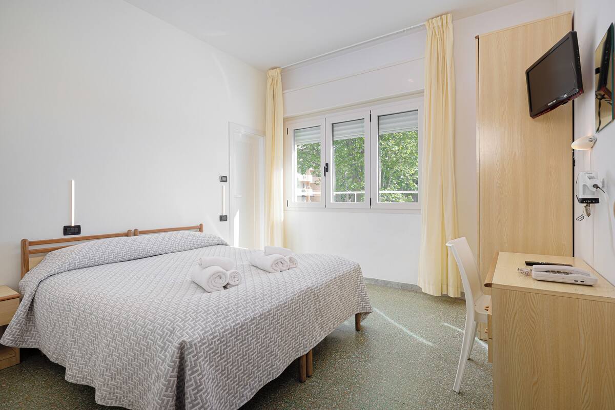 Habitación de hotel con cama, escritorio, silla, televisor y ventana con cortinas.