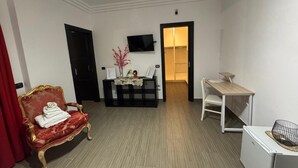 A hotel room with a red upholstered armchair, a small table with a vase of flowers, a desk with a chair, and a flat-screen TV mounted on the wall. - Bed and Breakfast Simonelli RoomS (Rocca Priora)