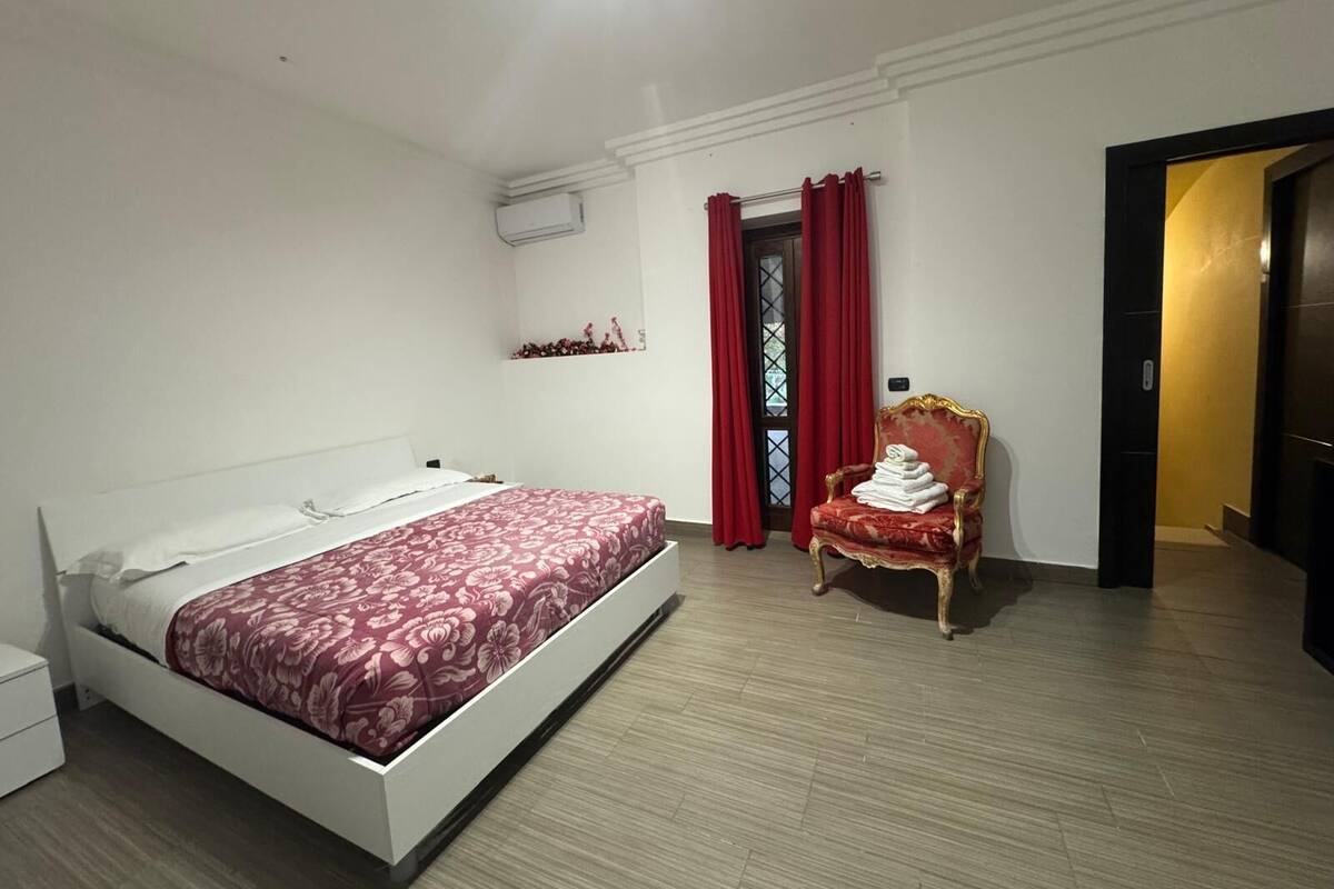 A bedroom with a bed, a chair, a window with red curtains, and a door.