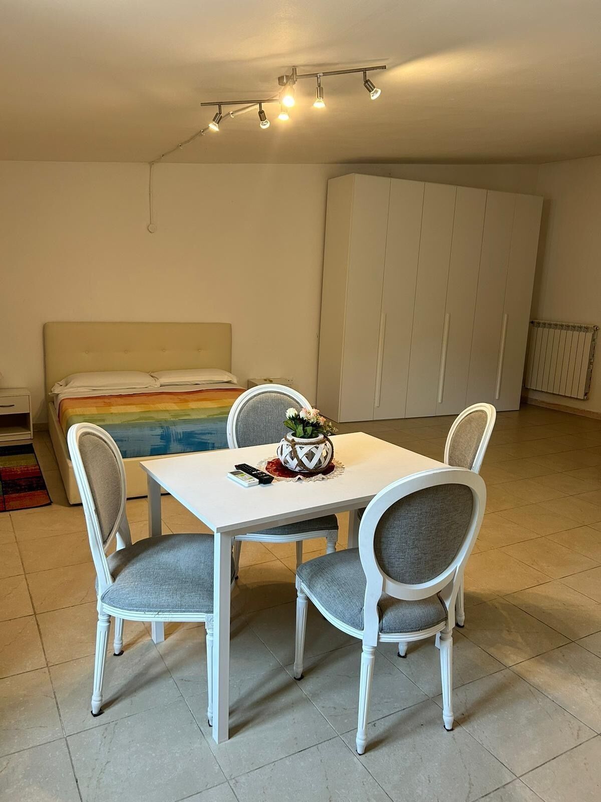 A room with a bed, a dining table with four chairs, a small table with a vase, and a ceiling light fixture.