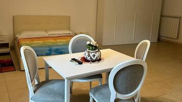 A room with a bed, a dining table with four chairs, a small table with a vase, and a ceiling light fixture.
