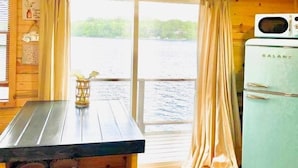 A cabin with a wooden interior, a dining table with plates, a refrigerator, and a view of a body of water through large windows.