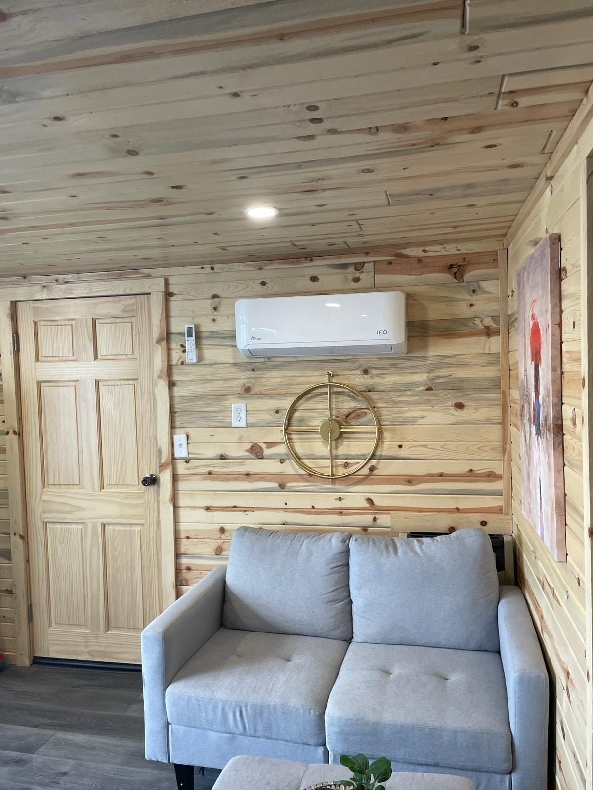 Ein Zimmer mit Holzboden, einem grauen Sofa, einer Wand-Klimaanlage und einer Tür.