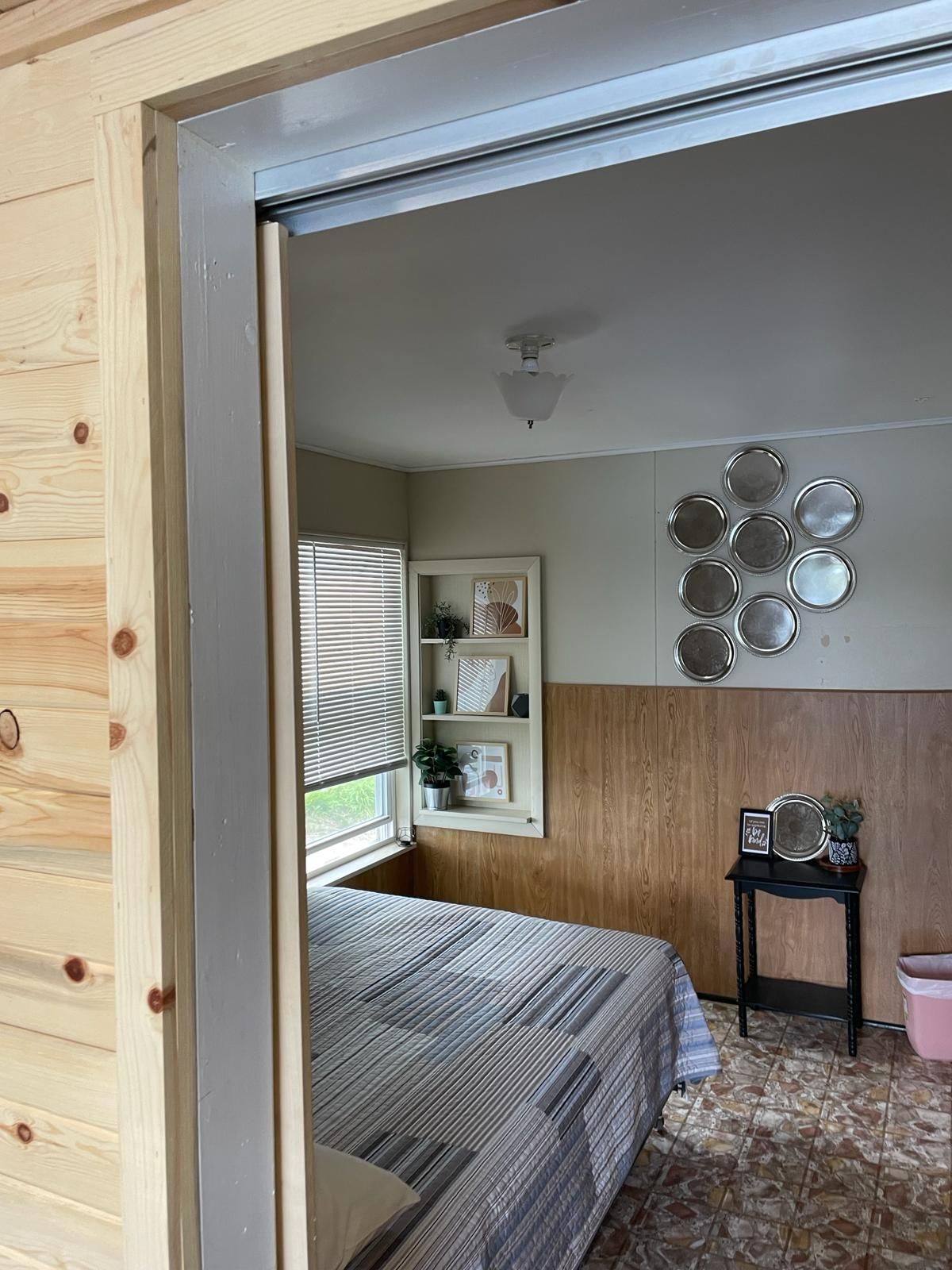 A bedroom with a bed, a window with blinds, a wall-mounted shelf with decorative items, and a ceiling light fixture.