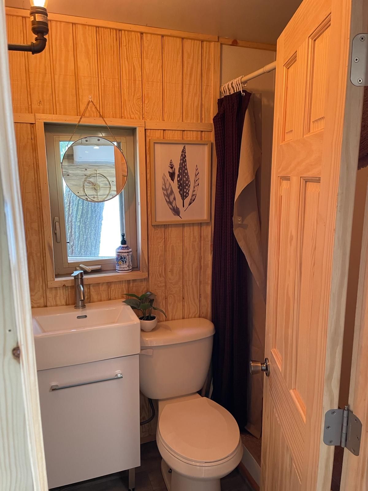 A compact bathroom with a wooden panel wall, a white sink, a toilet, a framed leaf artwork, and a window with a hanging light fixture.