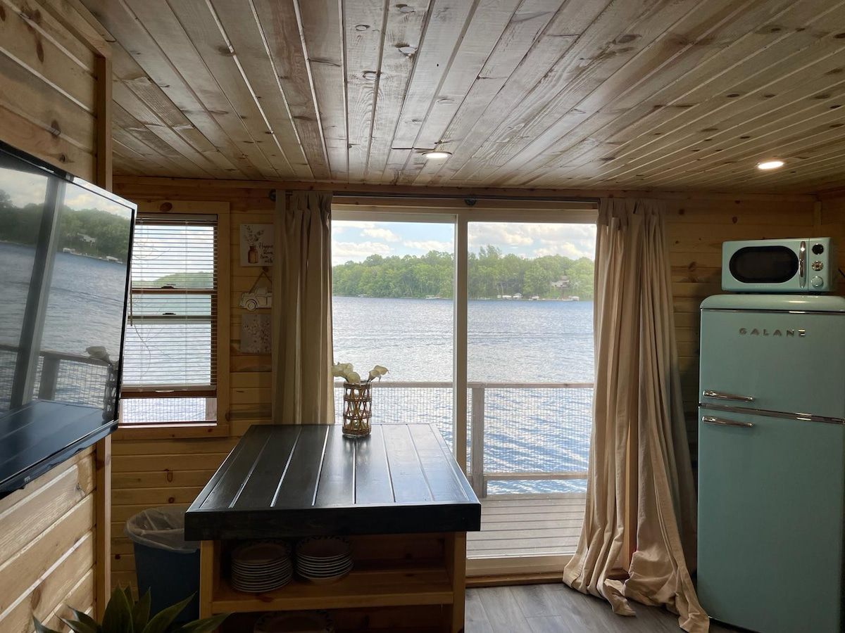 A cabin with a wooden interior, a dining table with a vase, a Galane refrigerator, and a view of a lake through a window.