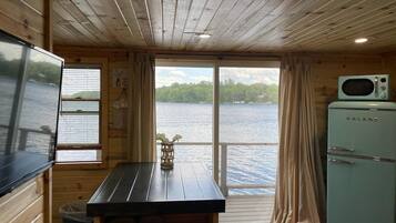 A cabin with a wooden interior, a dining table with a vase, a Galane refrigerator, and a view of a lake through a window.