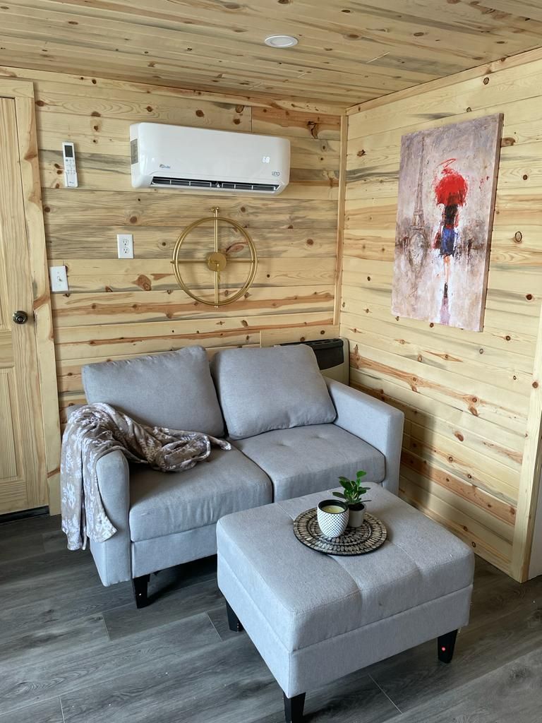 A room with a grey sofa, a matching ottoman, a wall-mounted air conditioner, and a painting.