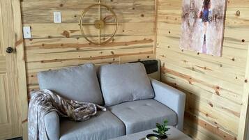 A room with a grey sofa, a matching ottoman, a wall-mounted air conditioner, and a painting.