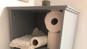 Ein grauer Kleiderschrank mit mehreren Regalen, der gefaltete Handtücher, eine Toilettenpapierrolle und ein schwarzes Objekt enthält.