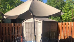 Eine Terrasse mit Tisch und Stühlen unter einem großen Sonnenschirm, umgeben von einem Holzzaun und Kübelpflanzen.