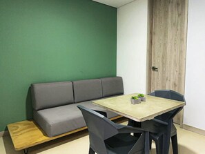 A room with a green wall, a grey sofa, a wooden bench, a table with two chairs, and a door with a wooden frame. - Moderno y Acogedor Apartamento Medellín Laureles (Medellín)