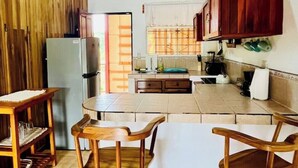 A kitchen with wooden cabinets, a refrigerator, a dining table with chairs, and a tiled floor. - Beautiful & Cozy Casa Aire, Near Lake Arenal (Nuevo Arenal)