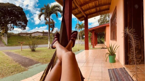A hammock under a wooden porch with a tiled floor, a potted plant, and a view of a road and houses in the background. - Beautiful & Cozy Casa Aire, Near Lake Arenal (Nuevo Arenal)
