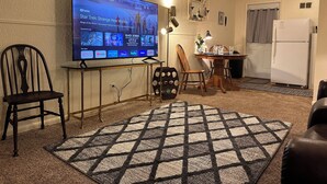 Een woonkamer met een flatscreen televisie op een standaard, een eettafel met stoelen, een keuken met een koelkast, en een kamerbreed tapijt met patroon.