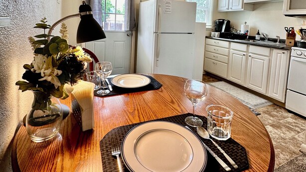 Een eettafel gedekt met borden, bestek en een bloevas in een goed verlichte keuken met witte kasten en een koelkast.