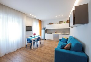 A modern living room with a blue sofa, a dining table with chairs, and a kitchen area with white cabinets and a built-in oven. - ELEGANT APARTMENT NEAR THE BEACH (Castellammare di Stabia)
