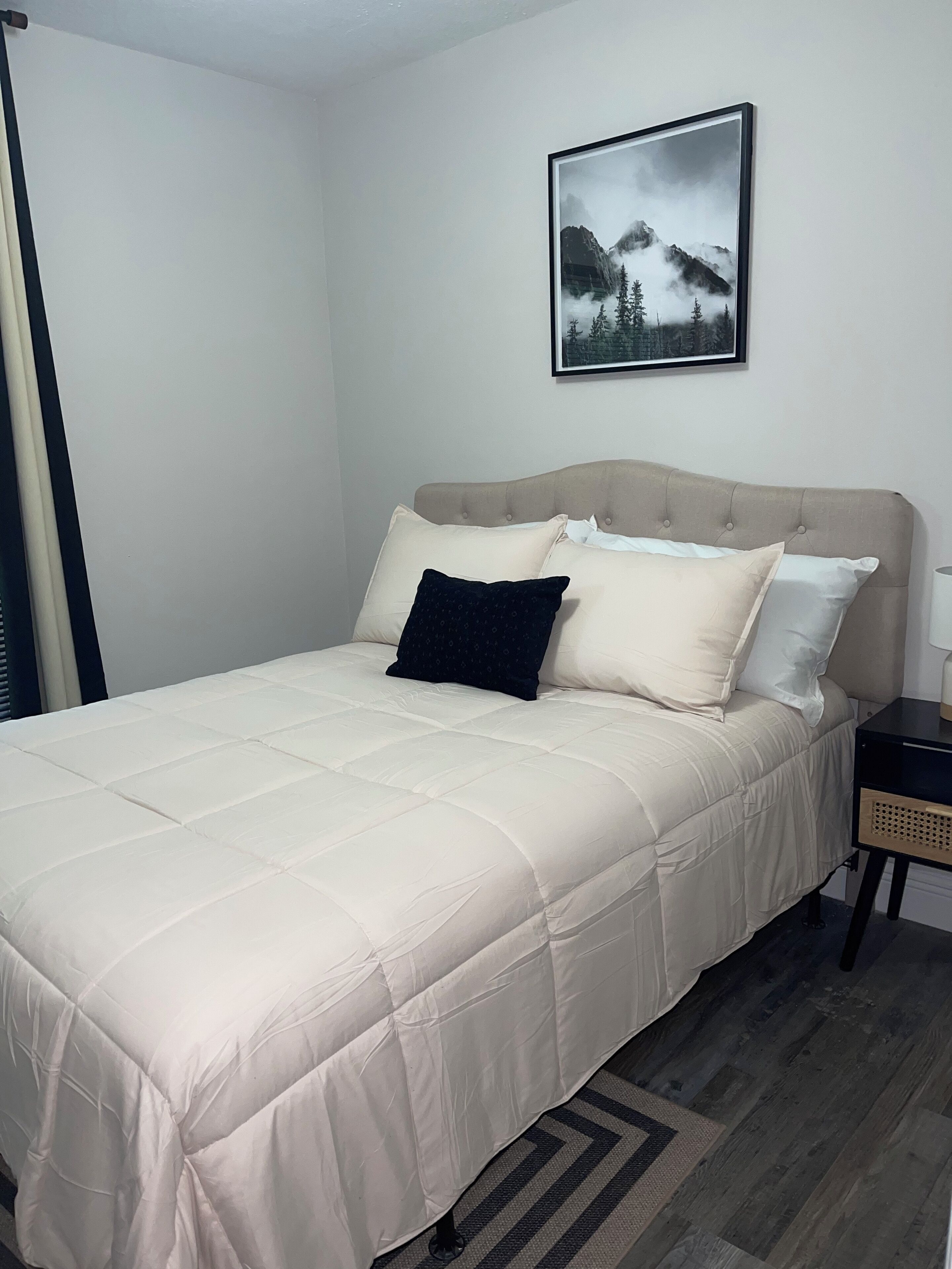 Un letto rifatto con cura, una testiera beige e biancheria da letto bianca, un quadro appeso alla parete e un comodino con una lampada.