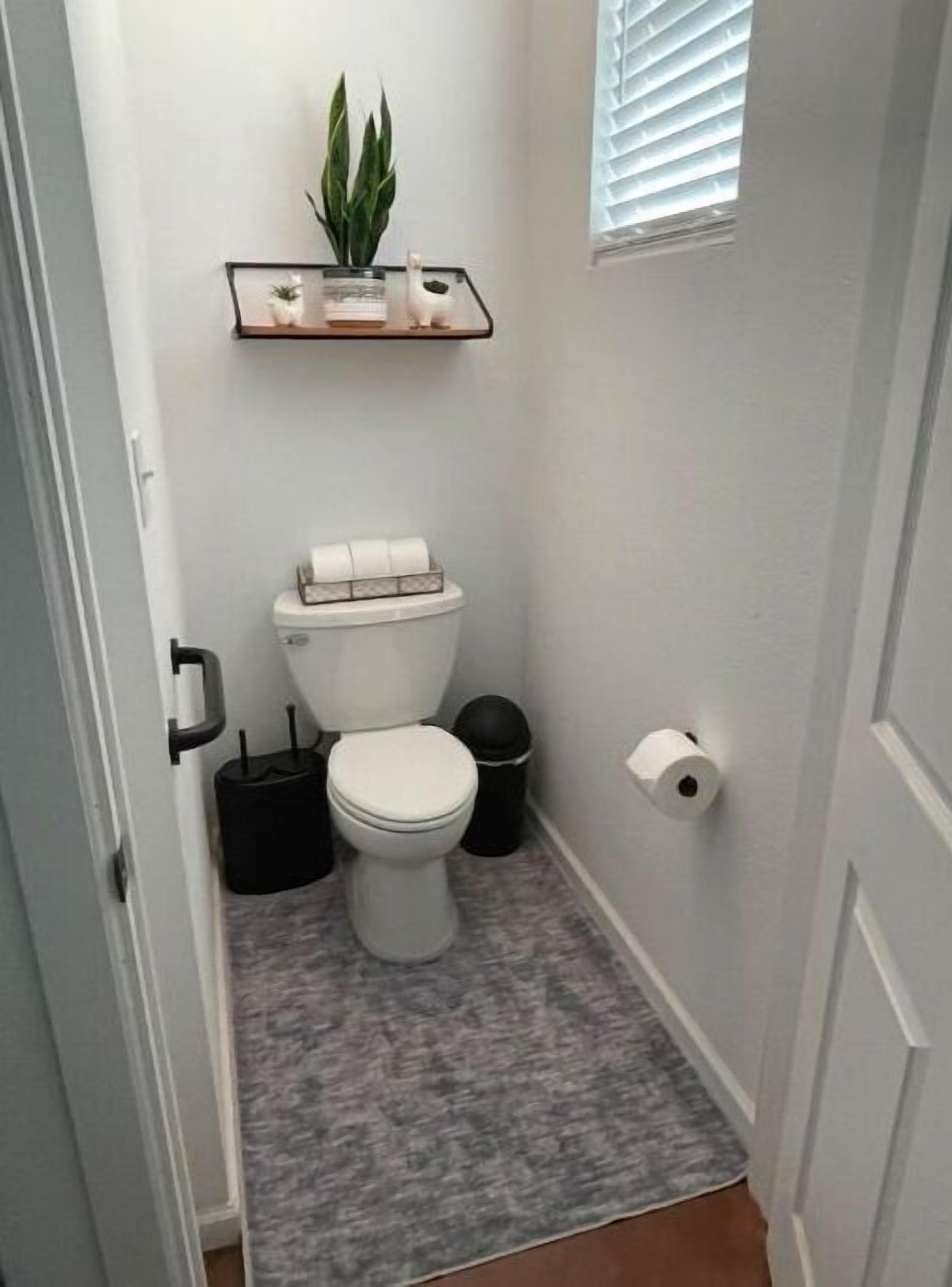 A small, narrow bathroom with a toilet, a trash can, a roll of toilet paper, and a potted plant on a shelf above the toilet.