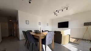 A modern living room with a dining area, a flat-screen TV mounted on the wall, and a large window allowing natural light. - Alba (Laxou)