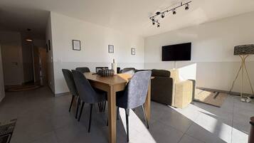 Una sala de estar moderna con comedor, un televisor de pantalla plana montado en la pared y un gran ventanal que deja entrar luz natural.