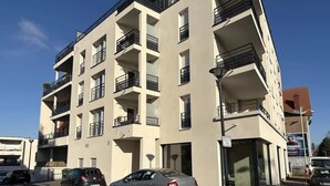 A multi-story building with balconies, a parking lot with cars, and a clear sky. - Alba (Laxou)